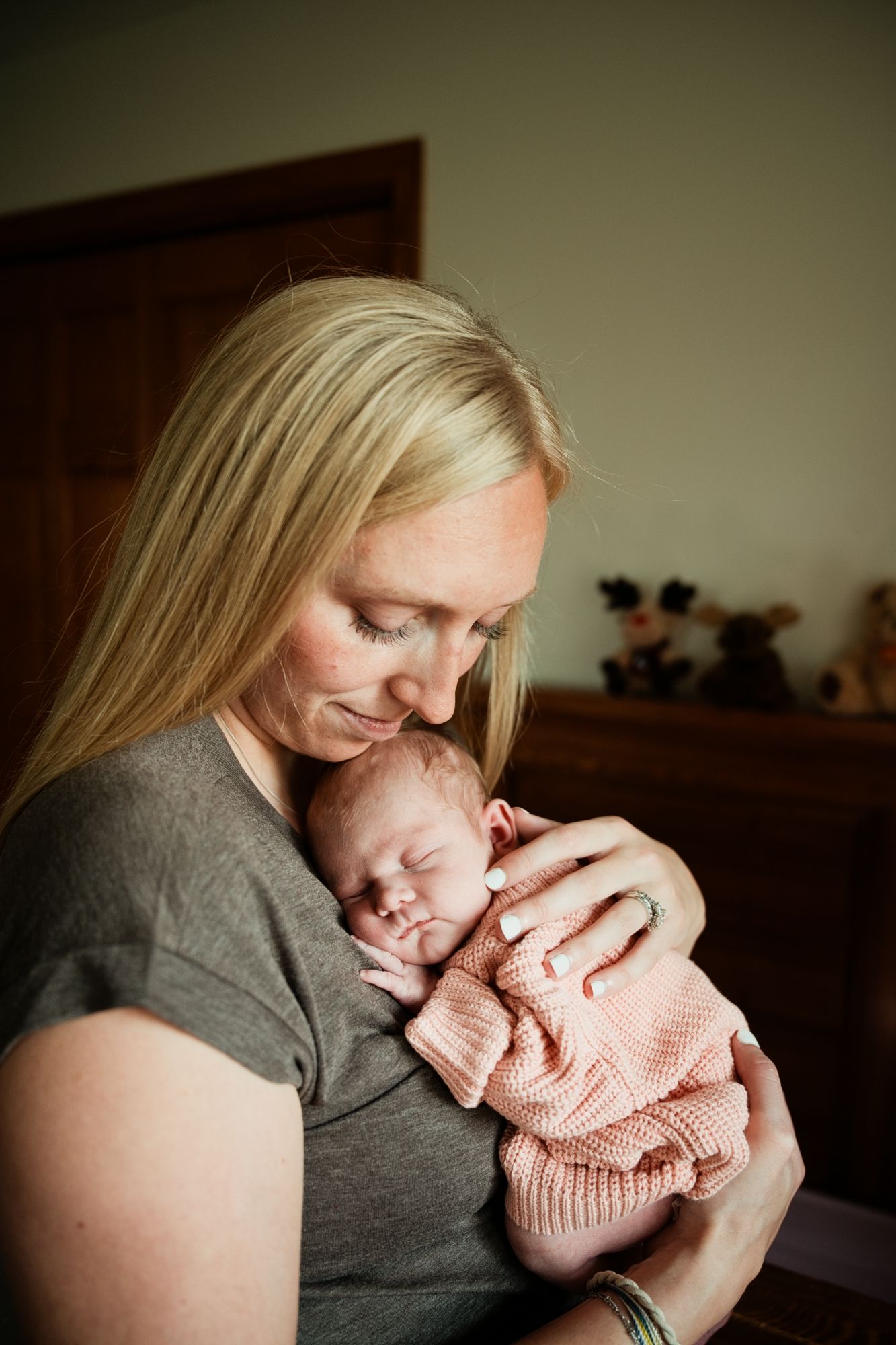 Blonde mother tenderly holding her sleeping newborn baby dressed in a pink knit outfit indoors.