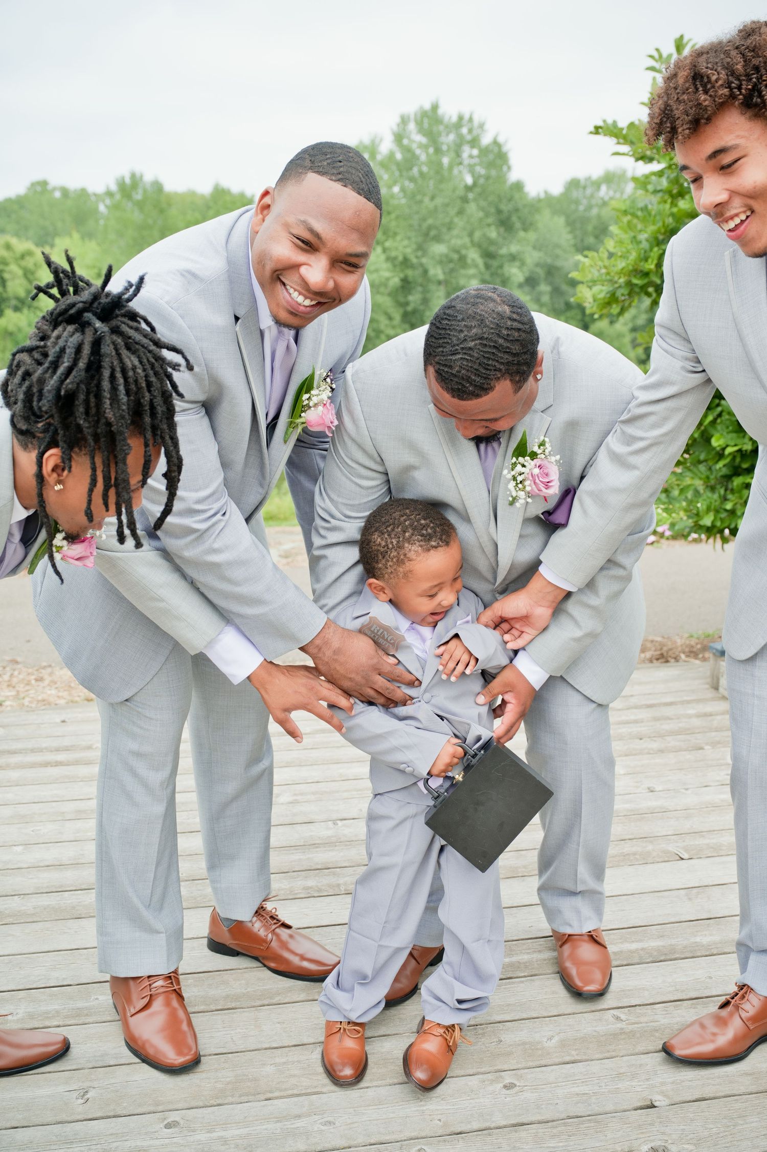 Groomsmen in gray suits laugh while helping young ring bearer on wooden deck.