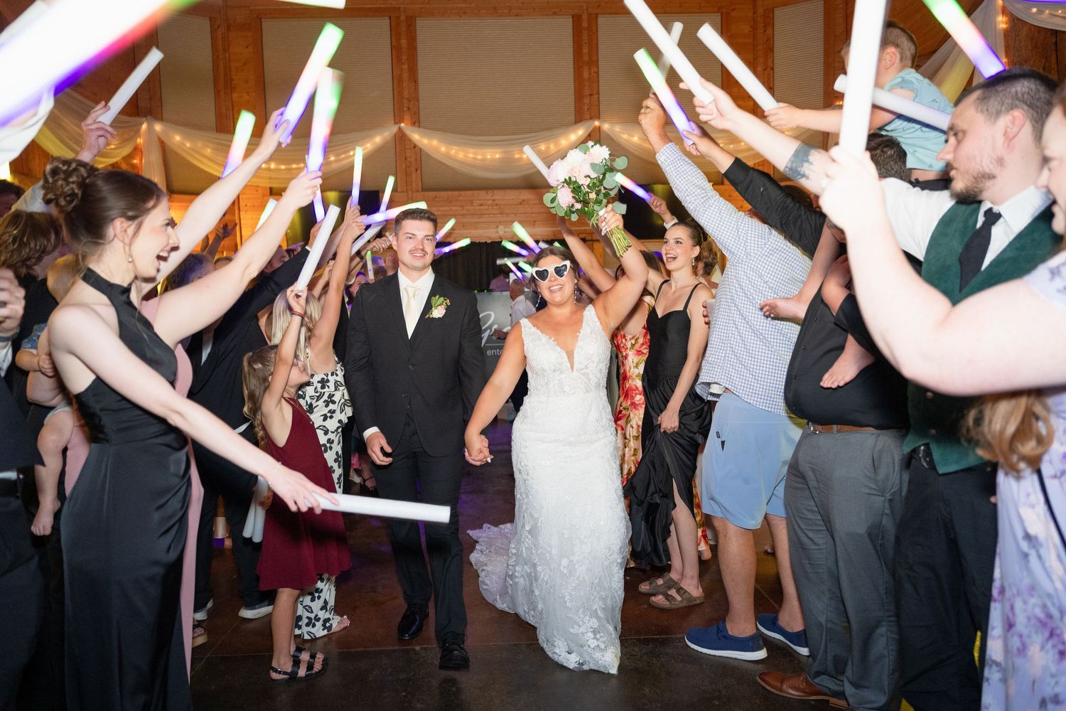 Bride and groom exit wedding reception through glowing foam stick tunnel held by cheering guests.