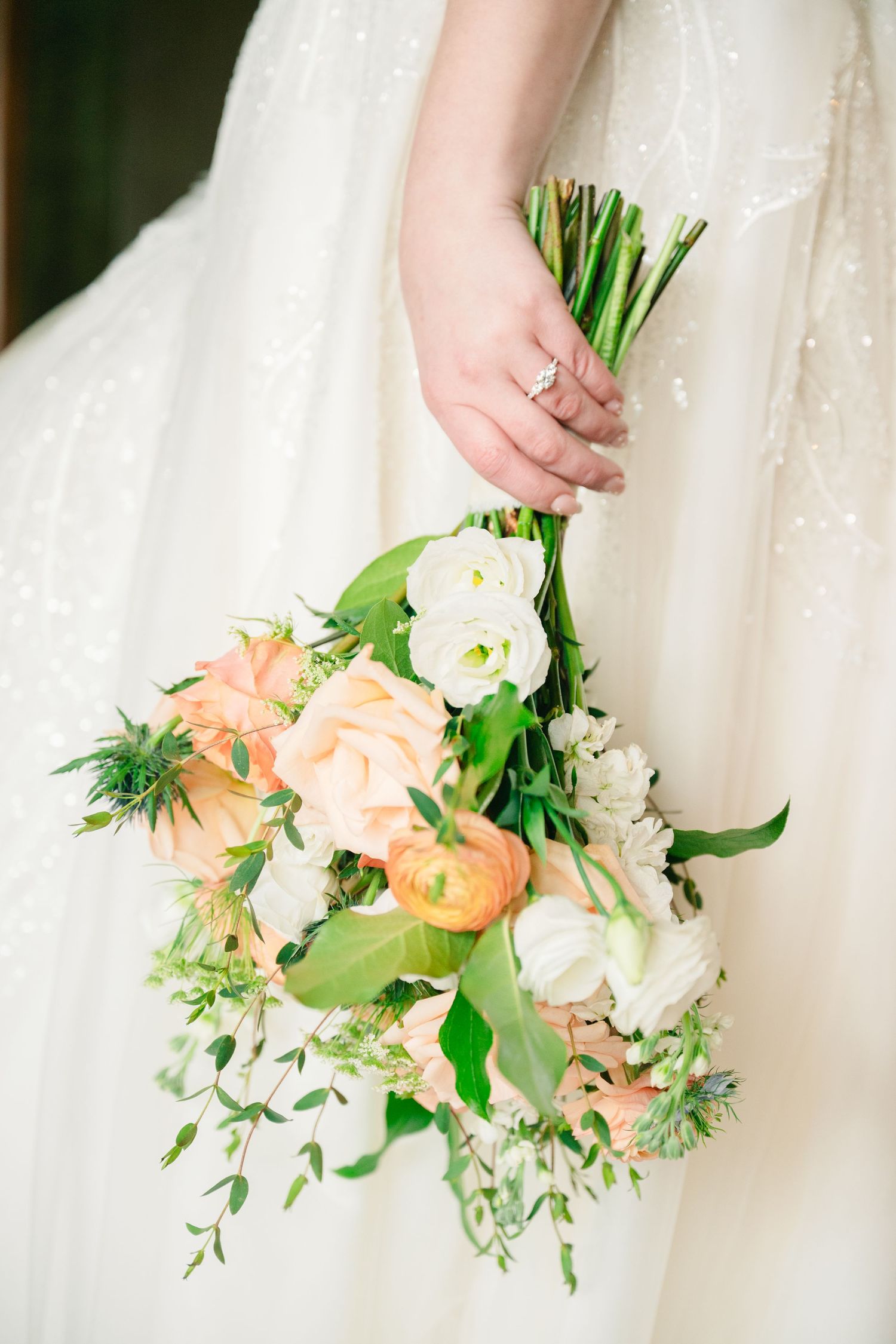 Bride holding a lush wedding bouquet with peach ranunculus, white roses, and greenery against a sparkly white gown.