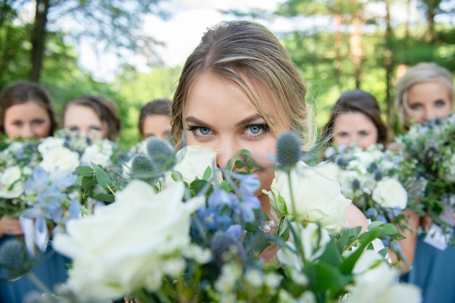 Creative photo of bridesmaids peeking over blue and white floral bouquets in outdoor garden setting. Zimmerman MN Weddings