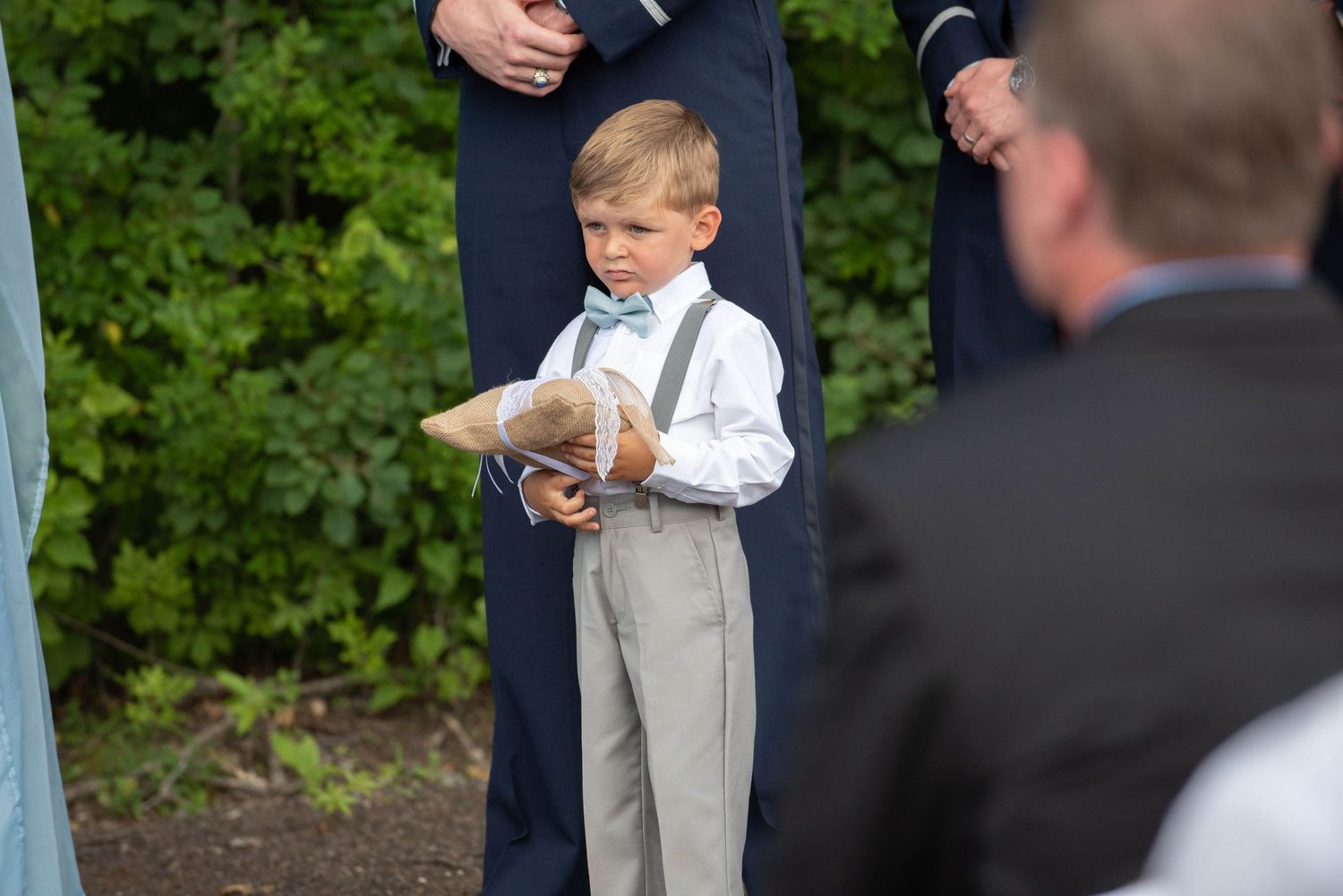 Young child stands between adults during an outdoor wedding ceremony with green foliage behind them. Zimmerman MN Weddings