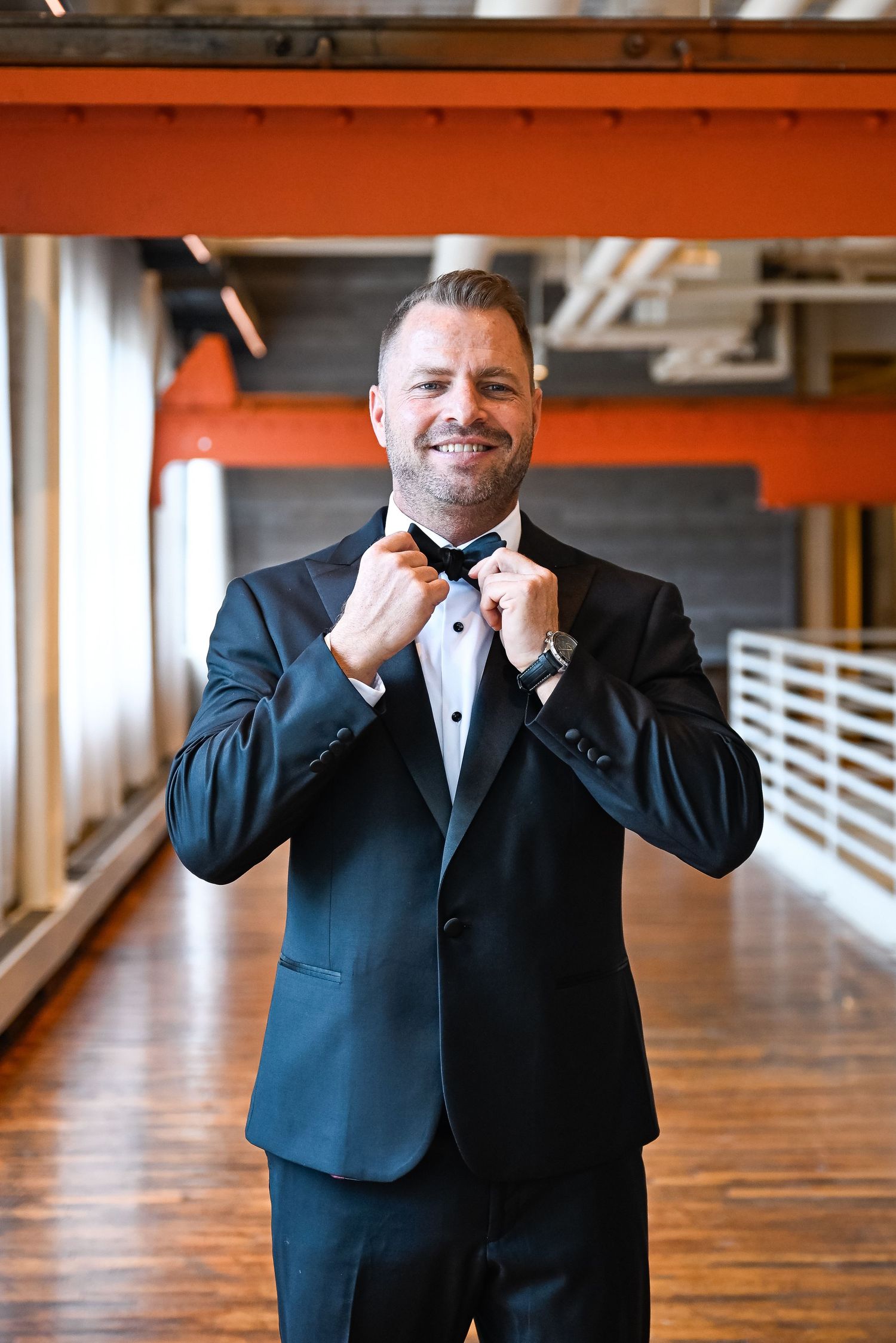 Person in tuxedo smiling and adjusting collar in industrial-style venue.