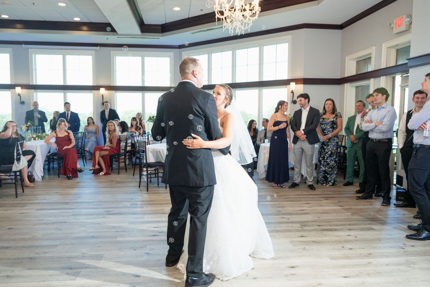 Bride and groom share first dance surrounded by guests in elegant reception hall with chandelier lighting.