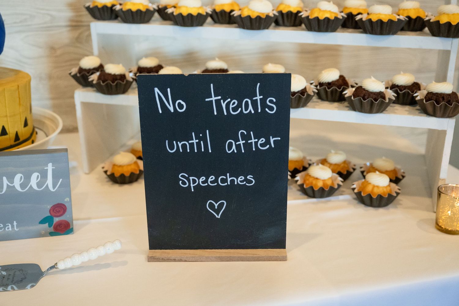 Chalkboard sign reading no treats until after speeches displayed on a dessert table with cupcakes.