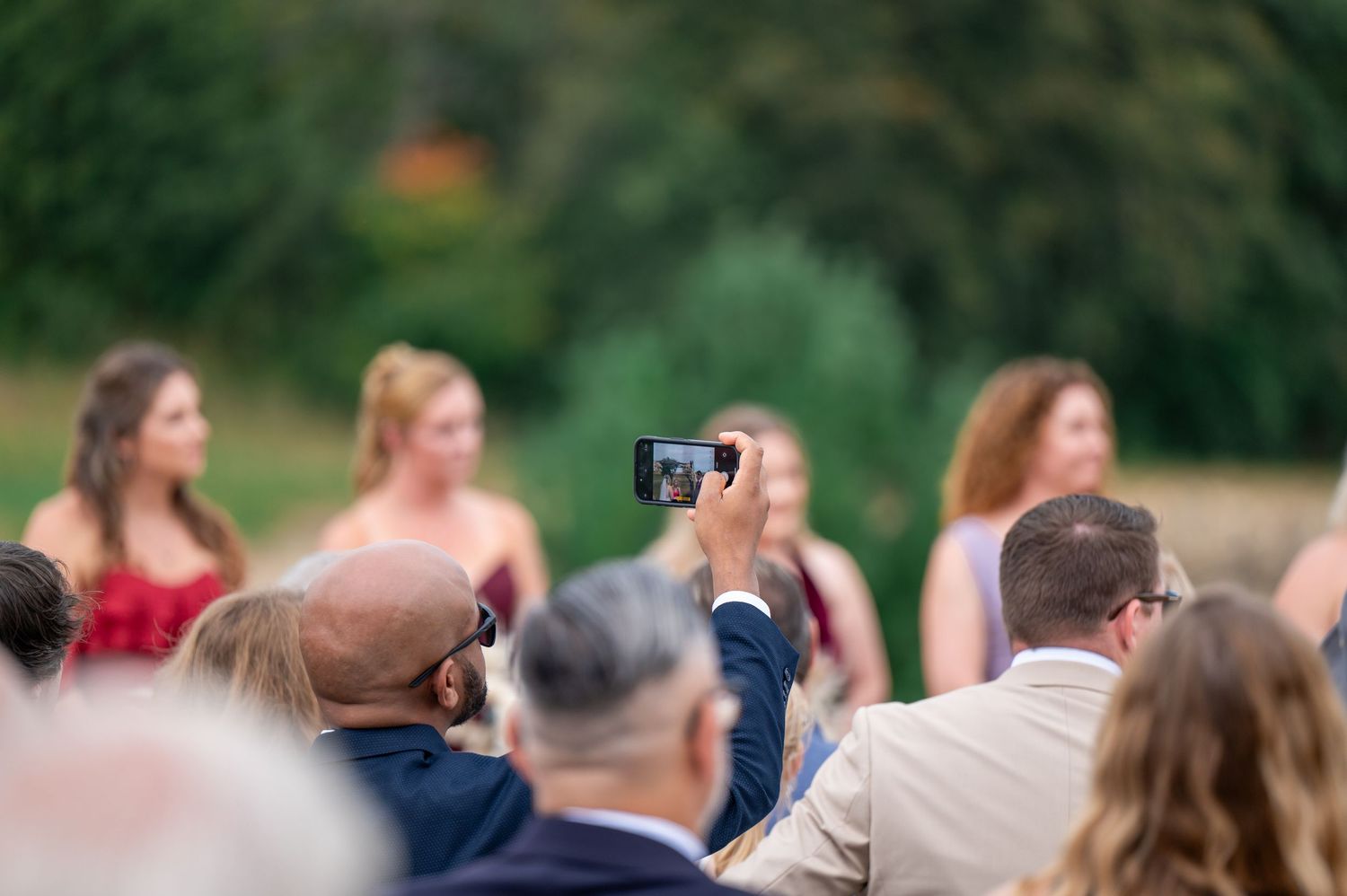 Guest captures wedding ceremony moments on smartphone among crowd of attendees.