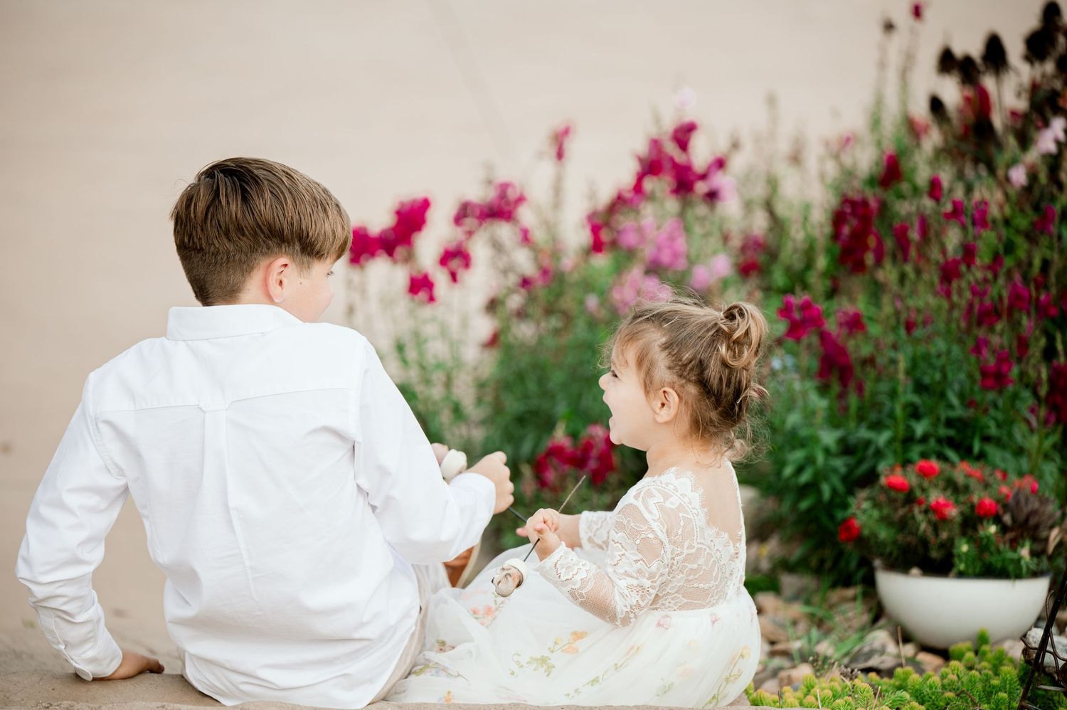 Two children sit together near vibrant pink flowers in formal wedding attire.