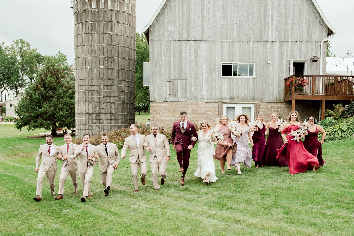 Joyful wedding party running on grass in front of white barn with burgundy and beige color scheme.
