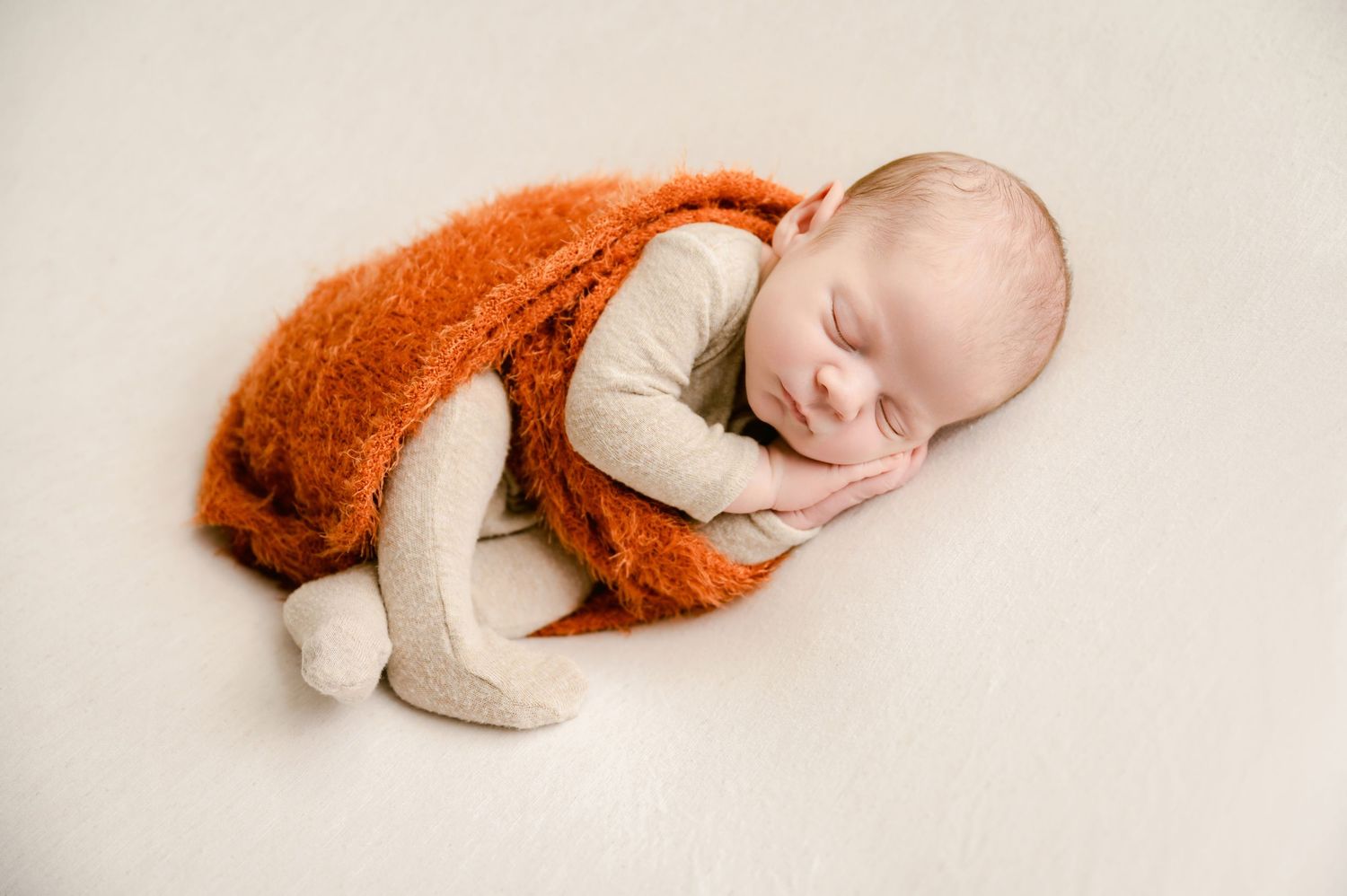 A sleeping baby in a white onesie lies curled up with a soft rust-colored blanket in a peaceful pose.