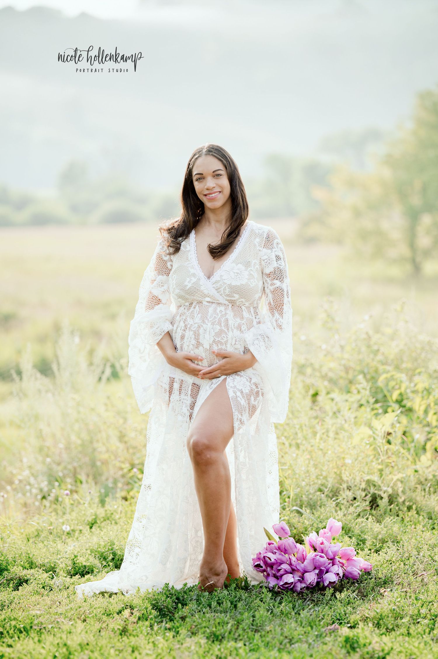 Pregnant woman in white lace maternity gown holding belly outdoors in field with purple flowers.