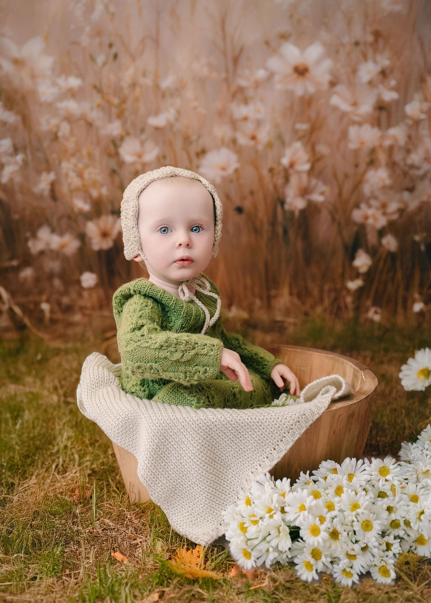 Adorable baby in green knit outfit and bonnet sitting in wooden bowl surrounded by white daisies and floral backdrop. mother day photoshoot