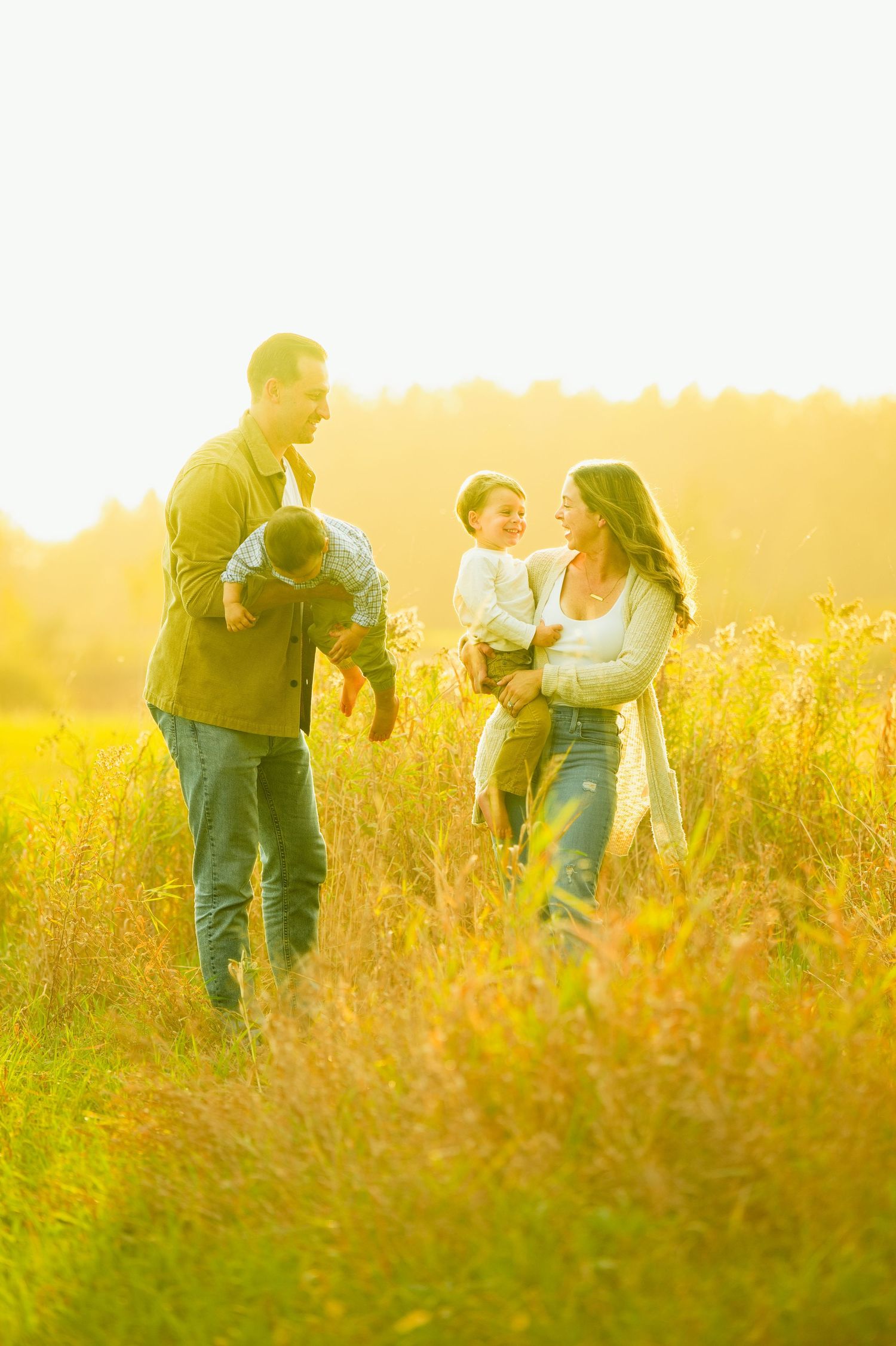 Family of four enjoying a sunny autumn day in a golden meadow, parents holding young children while smiling warmly.