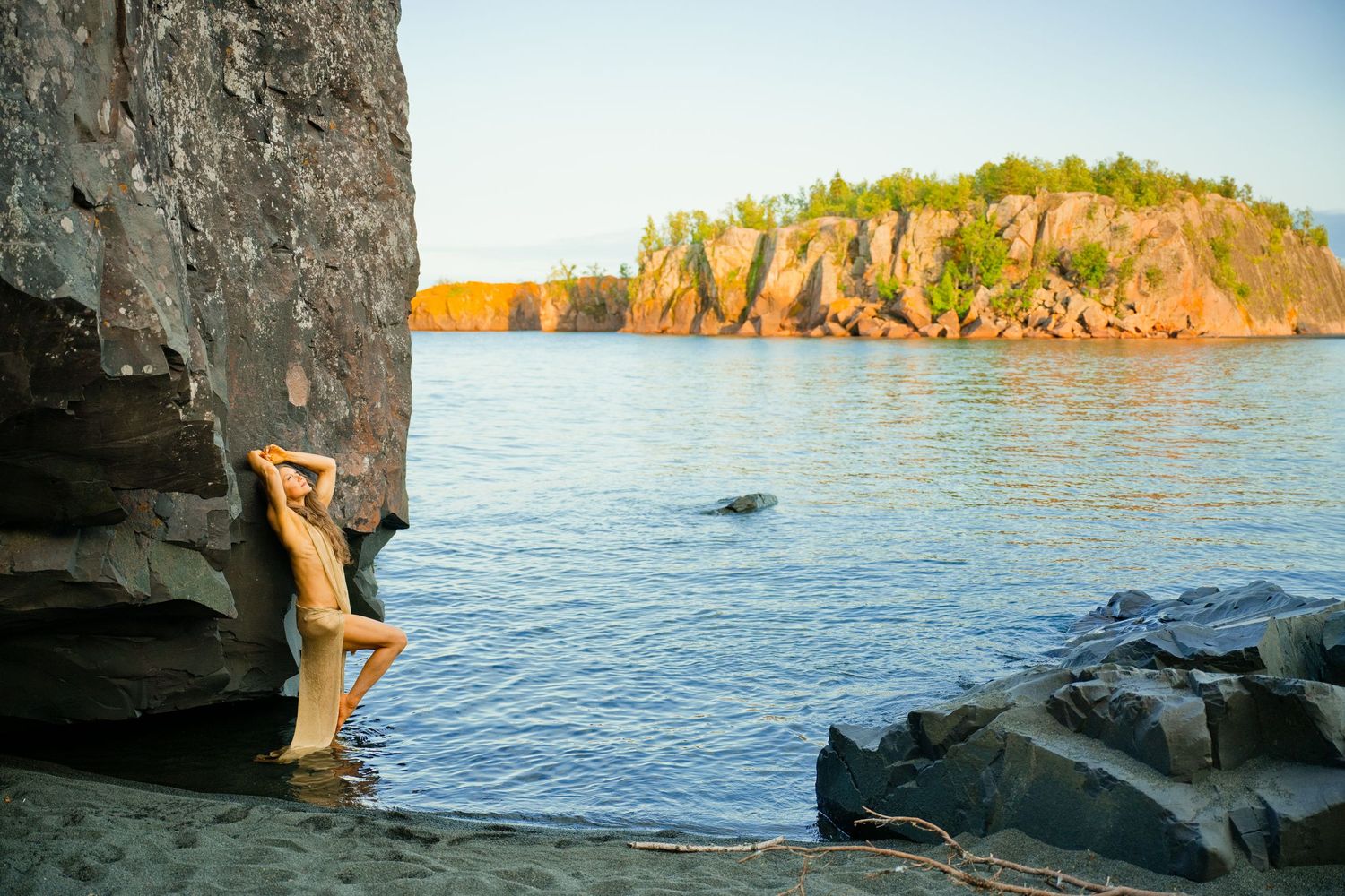 Person leaning against rocky cliff by calm lake with forested rock formations in golden sunlight.