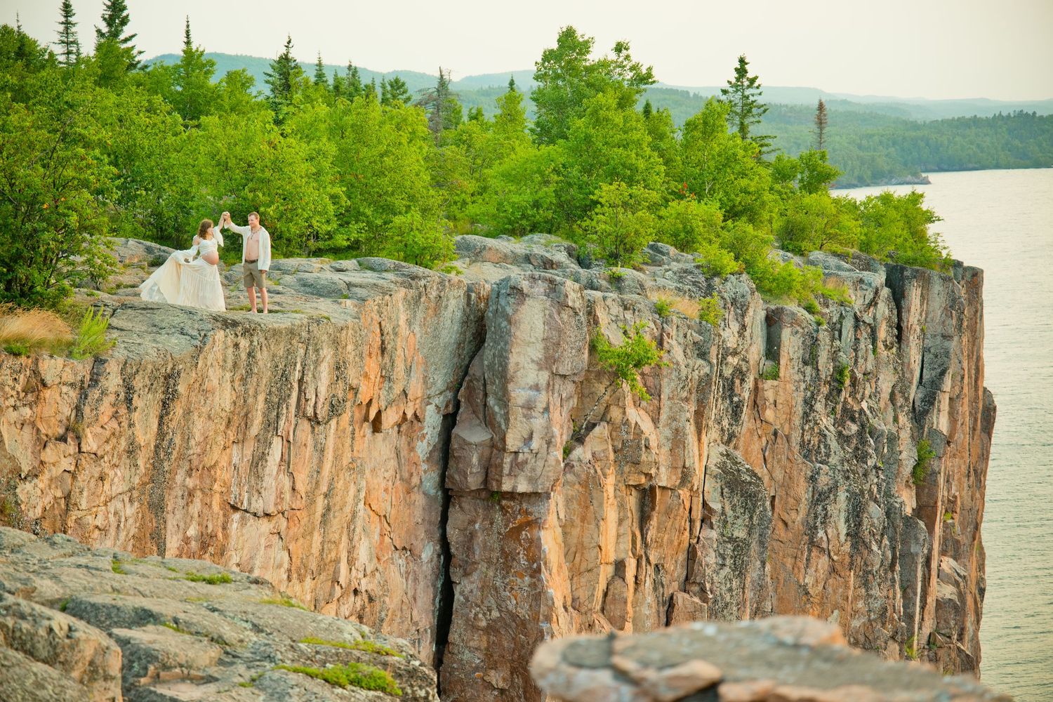 Couple standing on dramatic rocky cliffs overlooking a serene lake surrounded by lush green forest.