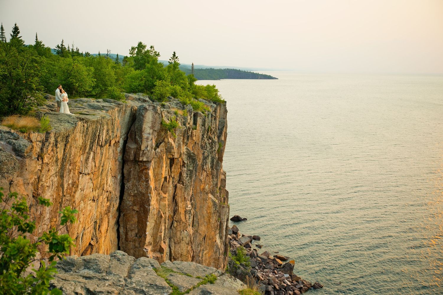 Couple stands atop dramatic rocky cliffs overlooking a vast lake surrounded by lush green trees at golden hour.