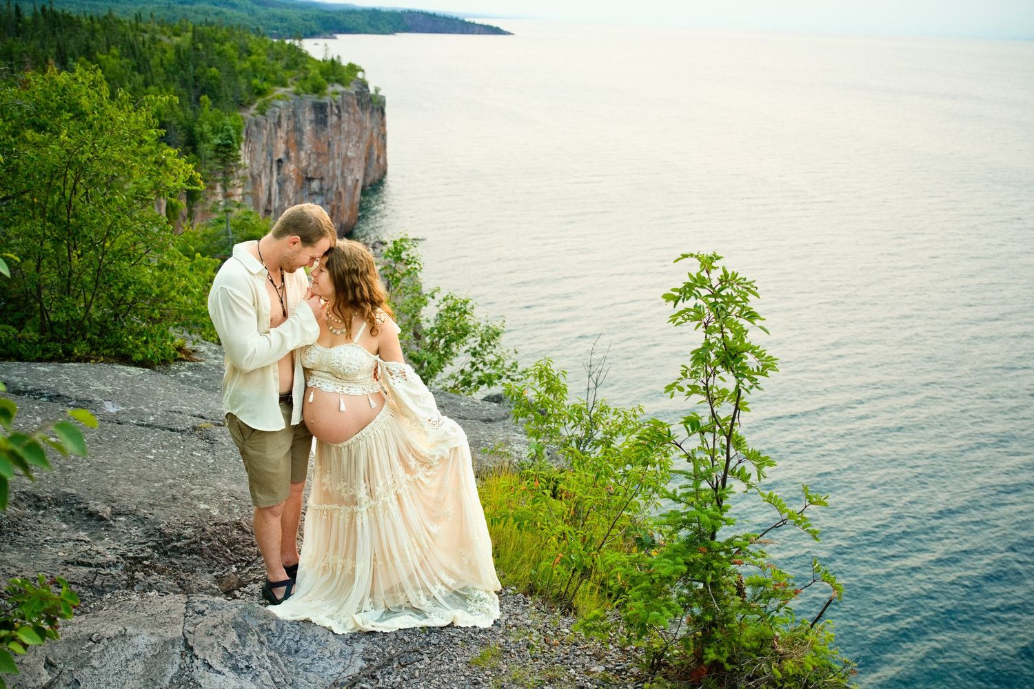 Pregnant couple sharing a romantic kiss on a cliff overlooking a serene lake, surrounded by lush greenery.