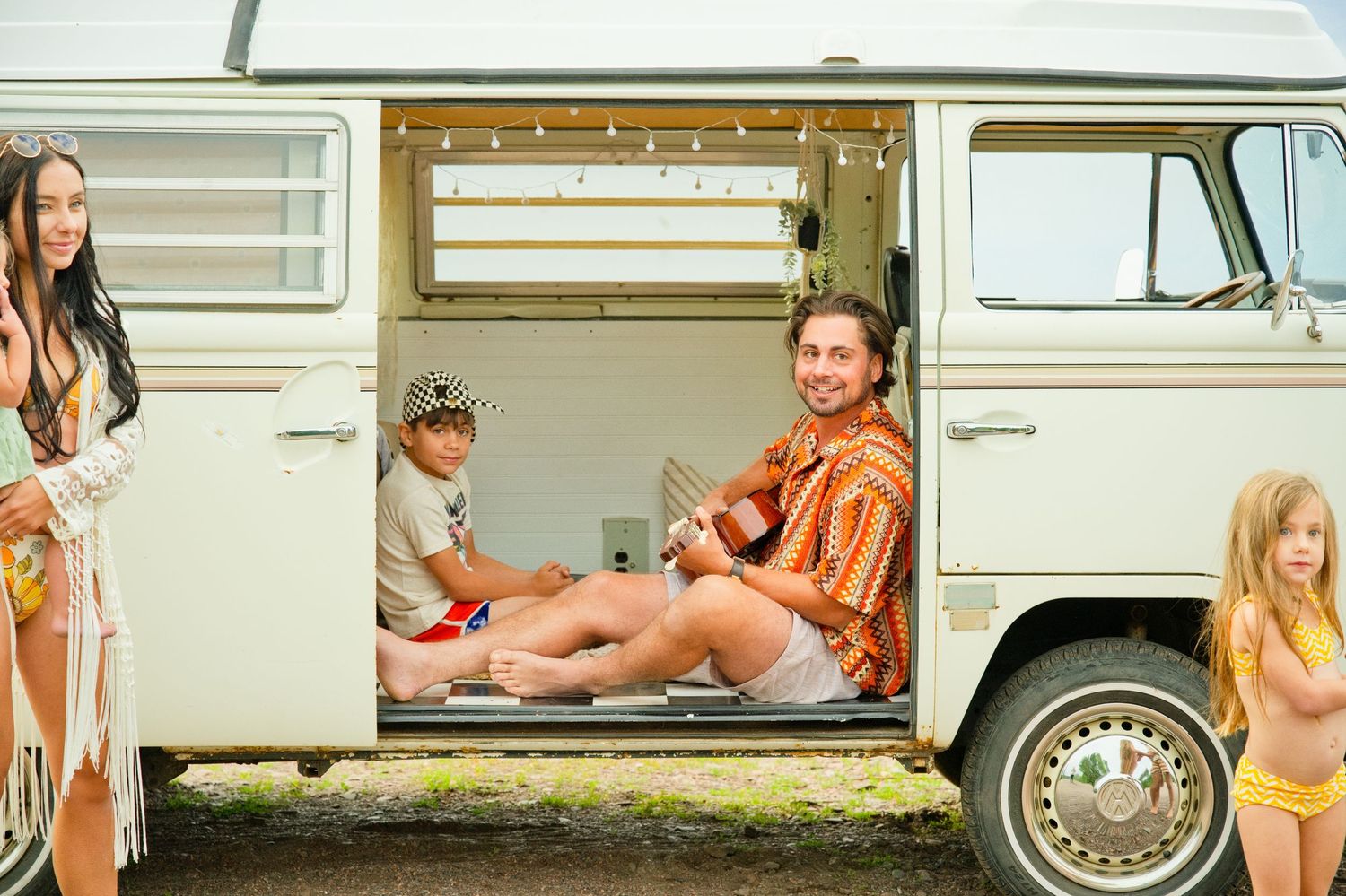 Family enjoying a vintage VW camper van on a sunny beach day, with kids and a man playing ukulele inside.