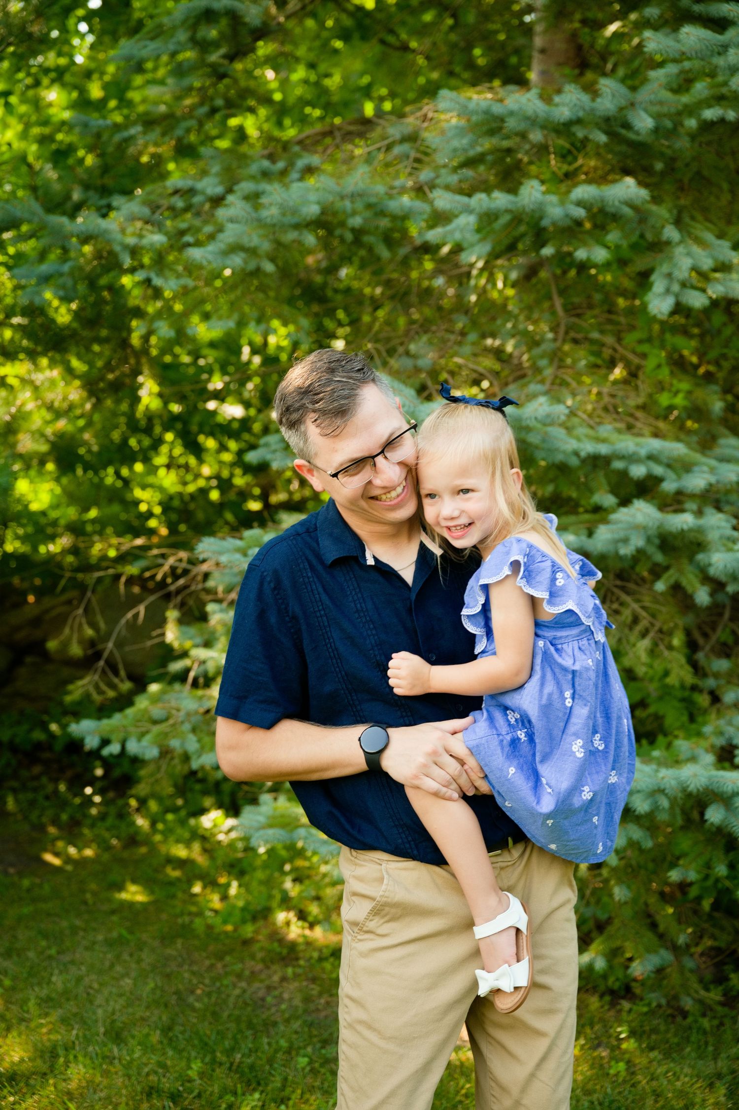 A smiling man in a navy shirt holds a laughing little girl in a blue dress outdoors among green trees.