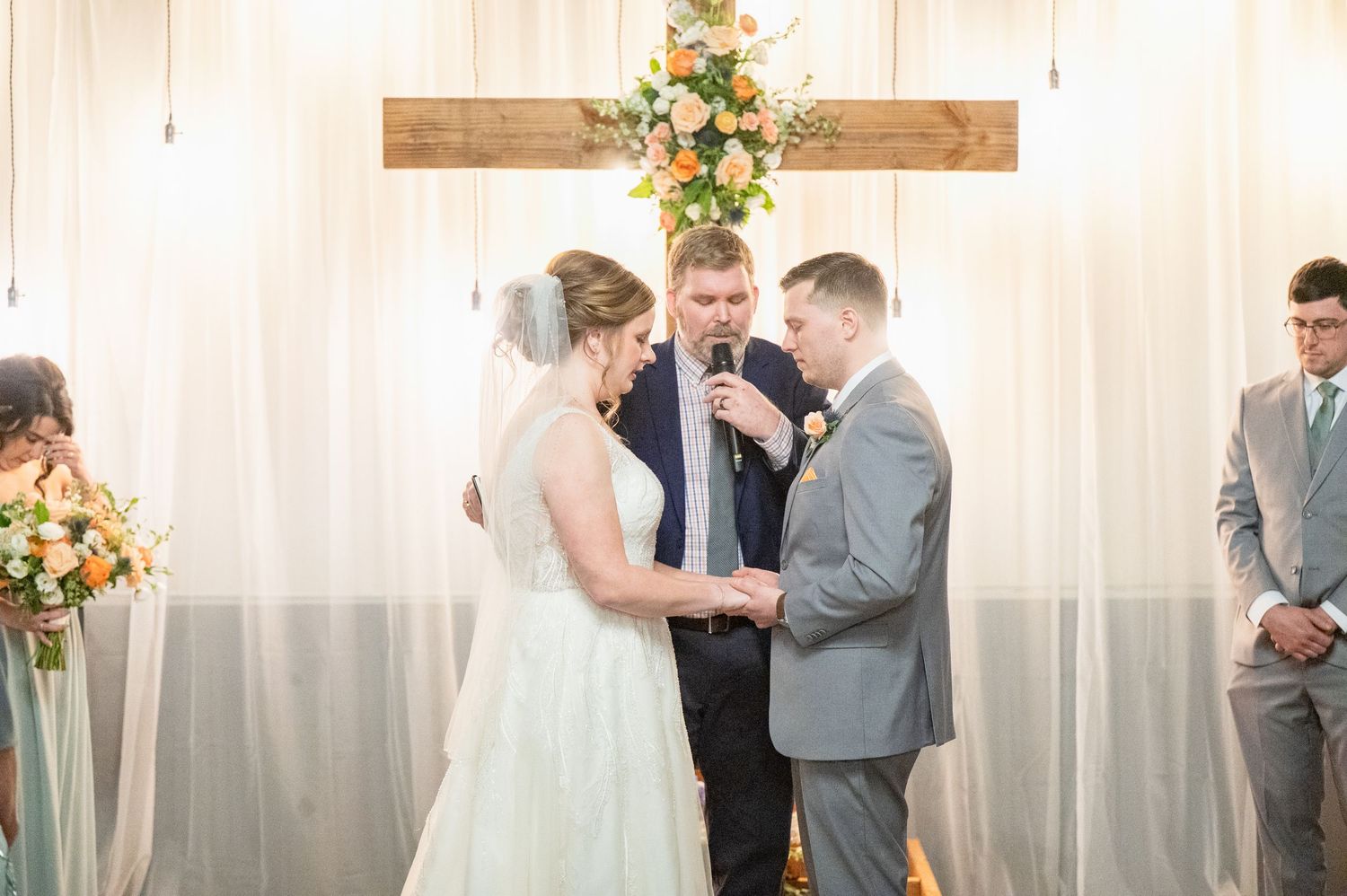 Bride and groom exchange vows with officiant at a decorated altar with floral arrangements and white draping.