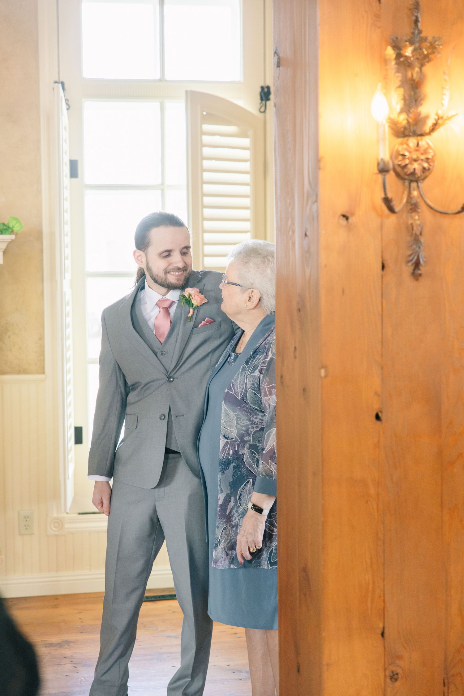 Groom in gray suit with pink tie shares a tender moment with elderly woman near wooden door at wedding venue.