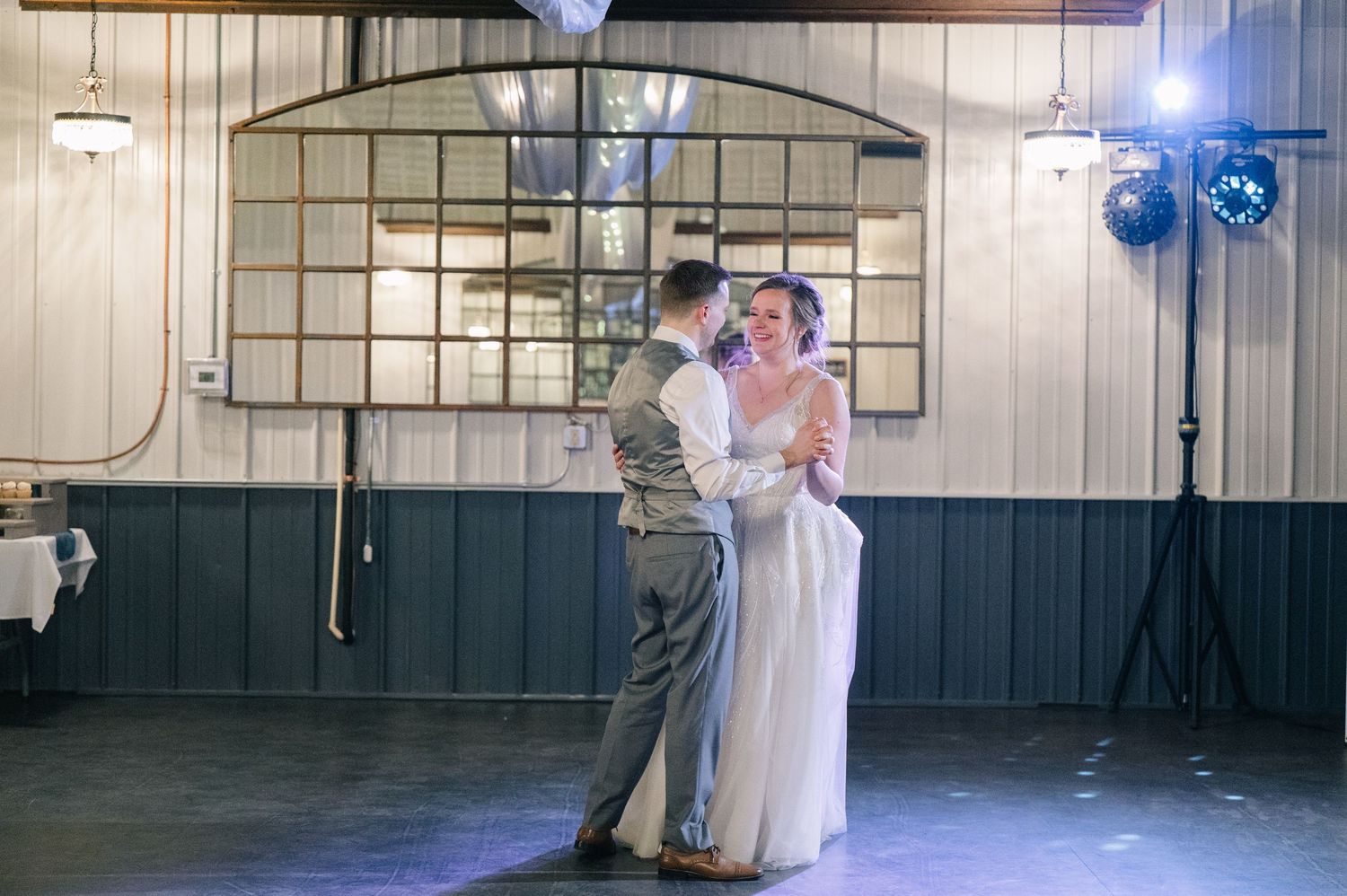 Bride and groom share romantic first dance in rustic wedding venue with industrial ceiling lights.