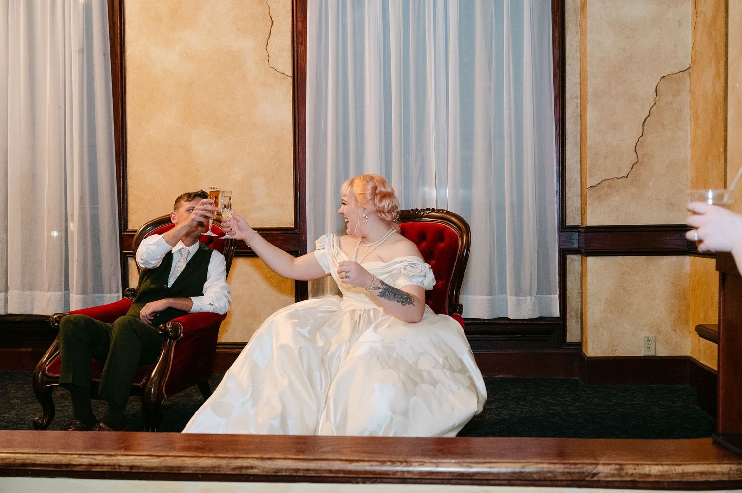 Central MN Wedding Photos | Bride and groom toasting with champagne glasses while seated in velvet chairs at wedding reception.