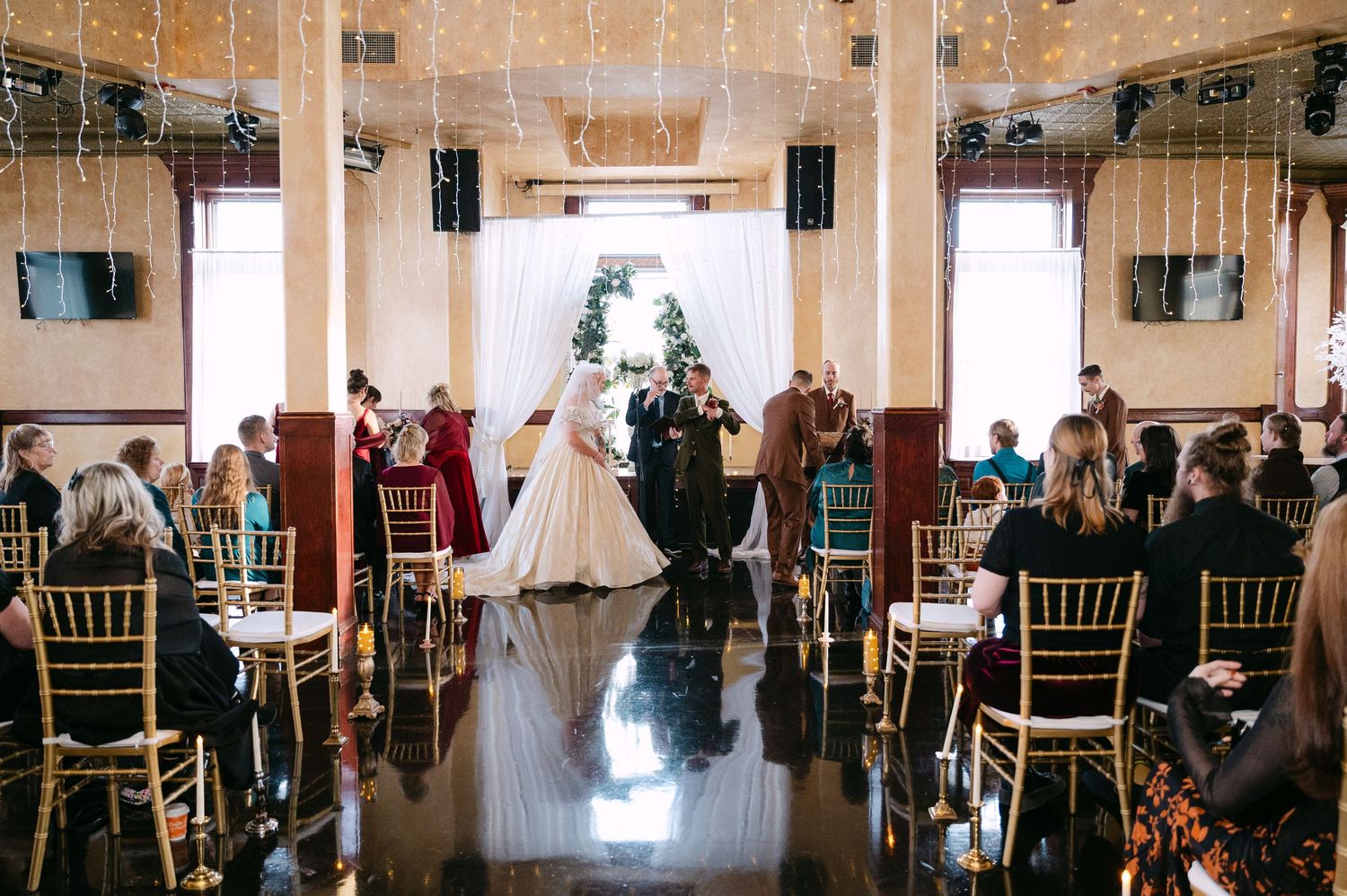 Central MN Wedding ceremony in elegant venue with string lights, guests seated in gold chairs as couple exchanges vows at floral arch.