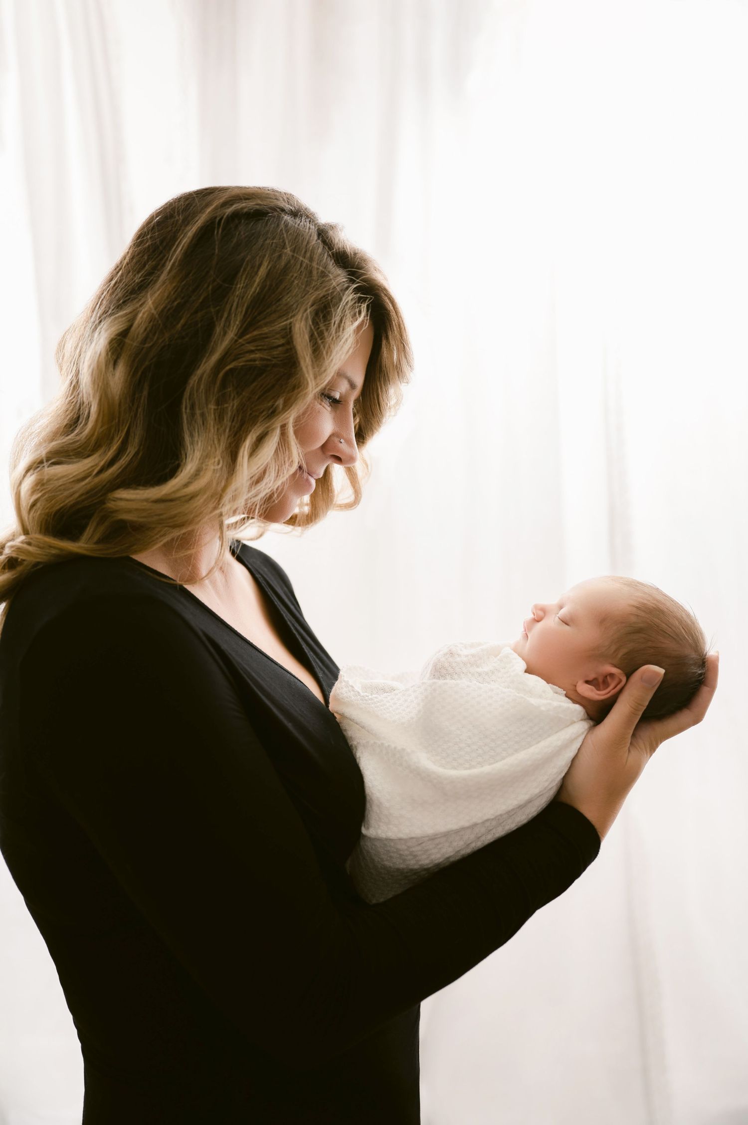 Central MN Baby Photos - A mother in a black dress tenderly holds her sleeping newborn baby wrapped in white, backlit by soft window light.