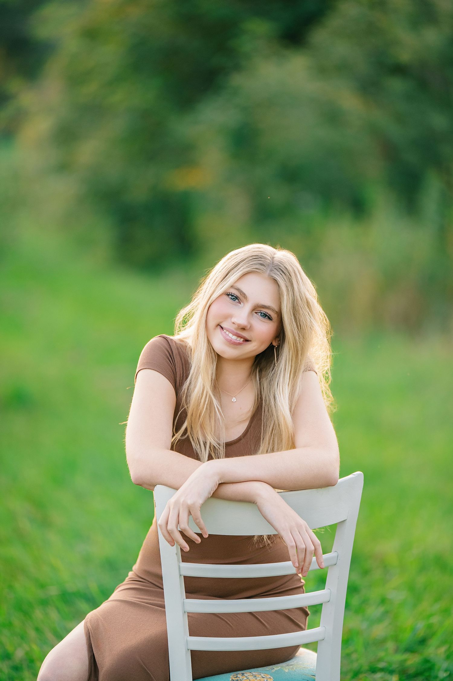 Central MN Senior Photography - Smiling blonde woman in brown dress sitting on white chair outdoors on green grass with trees in background.