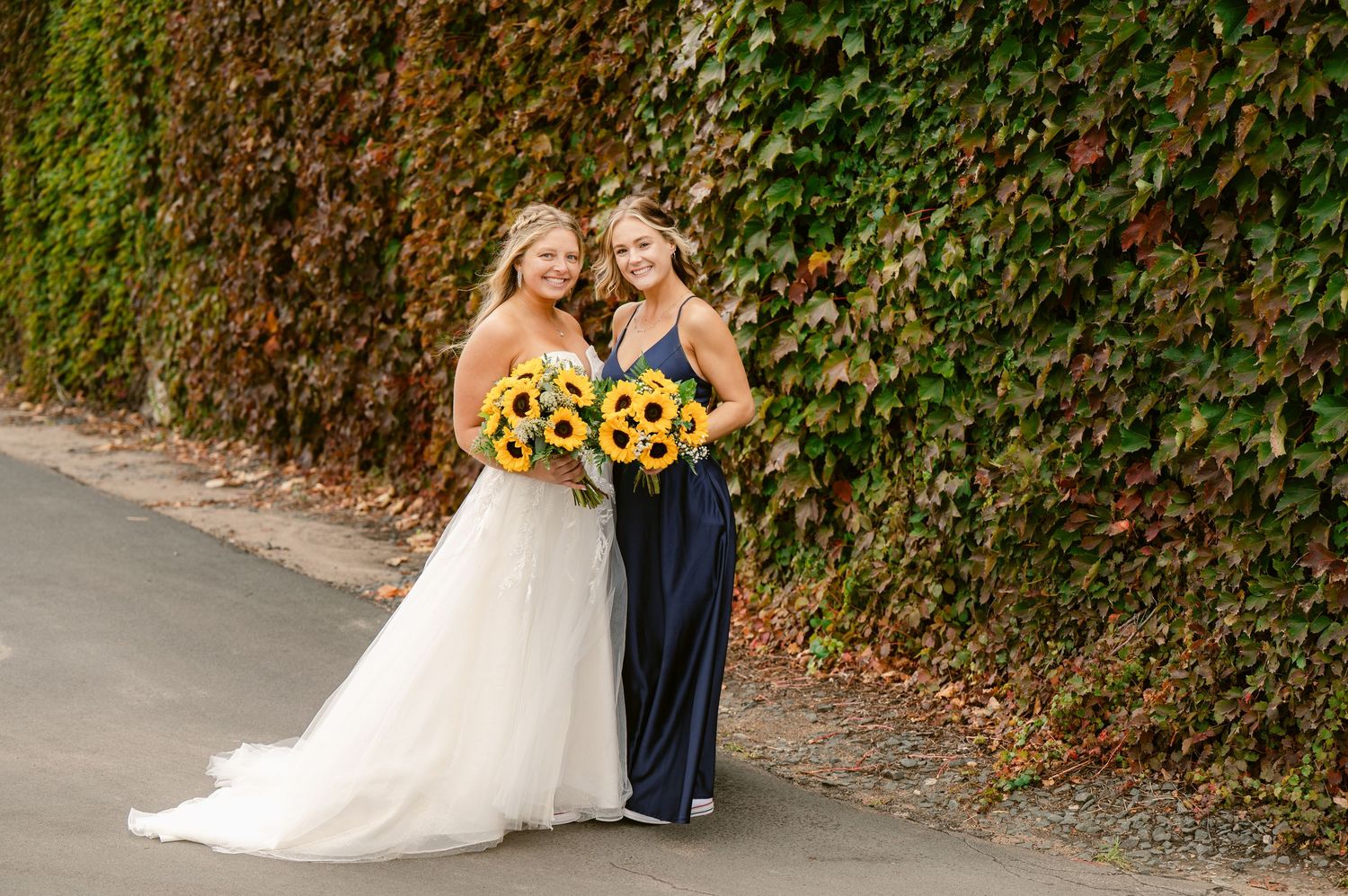 Central MN Wedding Photos | Bride and bridesmaid holding sunflower bouquets smile together in front of an ivy-covered wall.