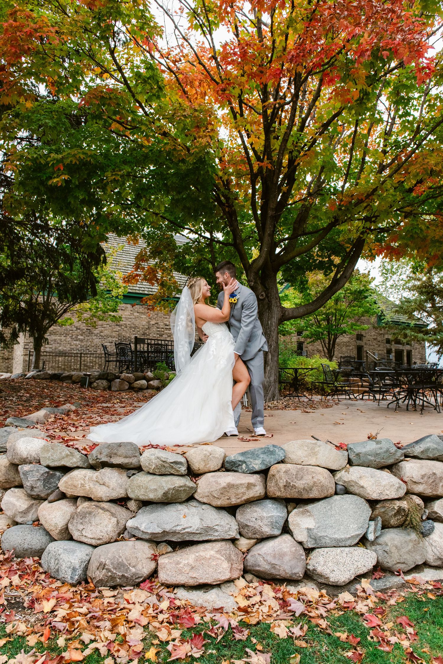 Central MN Wedding Photos | Bride and groom embrace on a stone wall surrounded by vibrant fall foliage and trees.