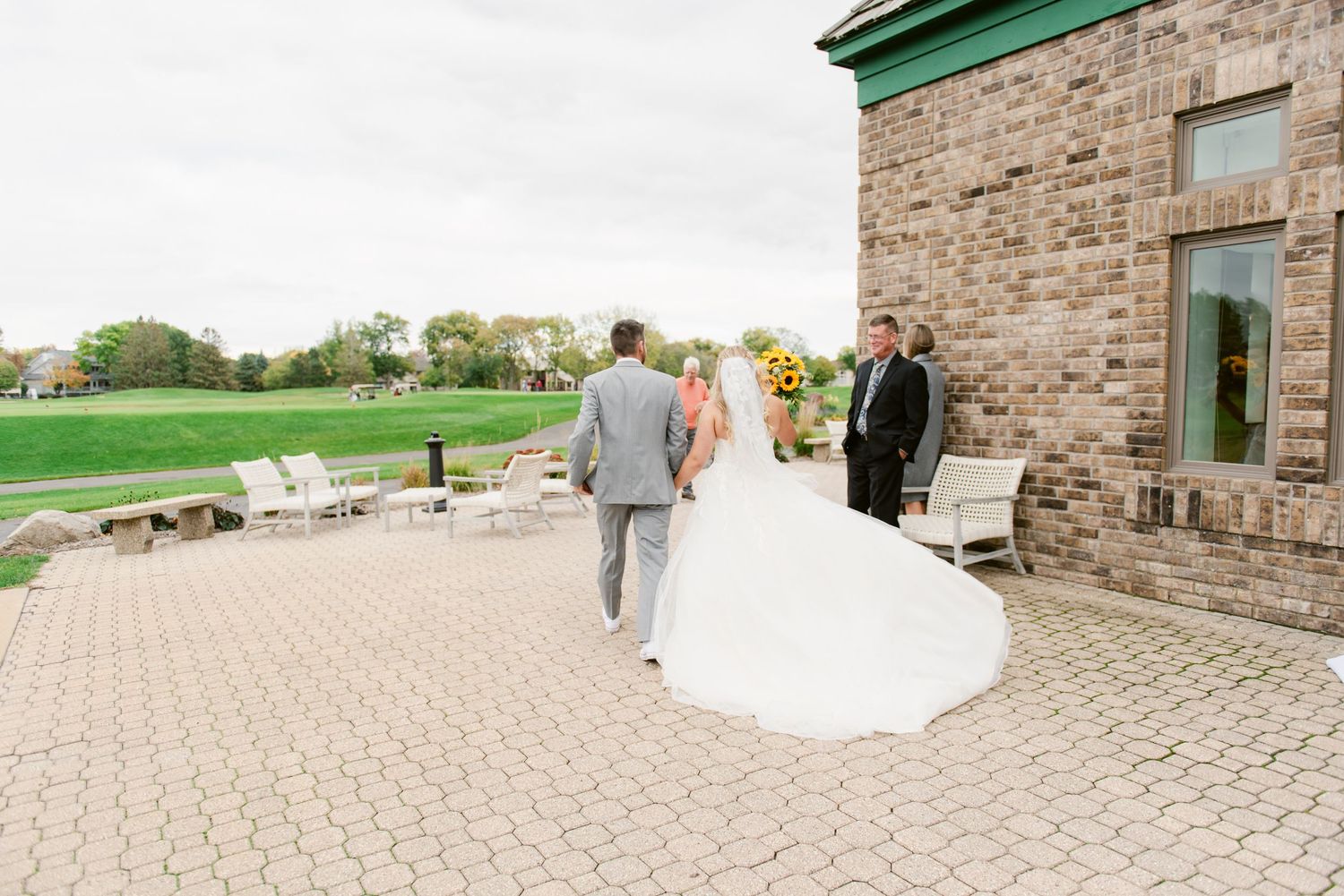 Central MN Wedding Photos - Bride in white gown with long train walking toward wedding officiant on patio.