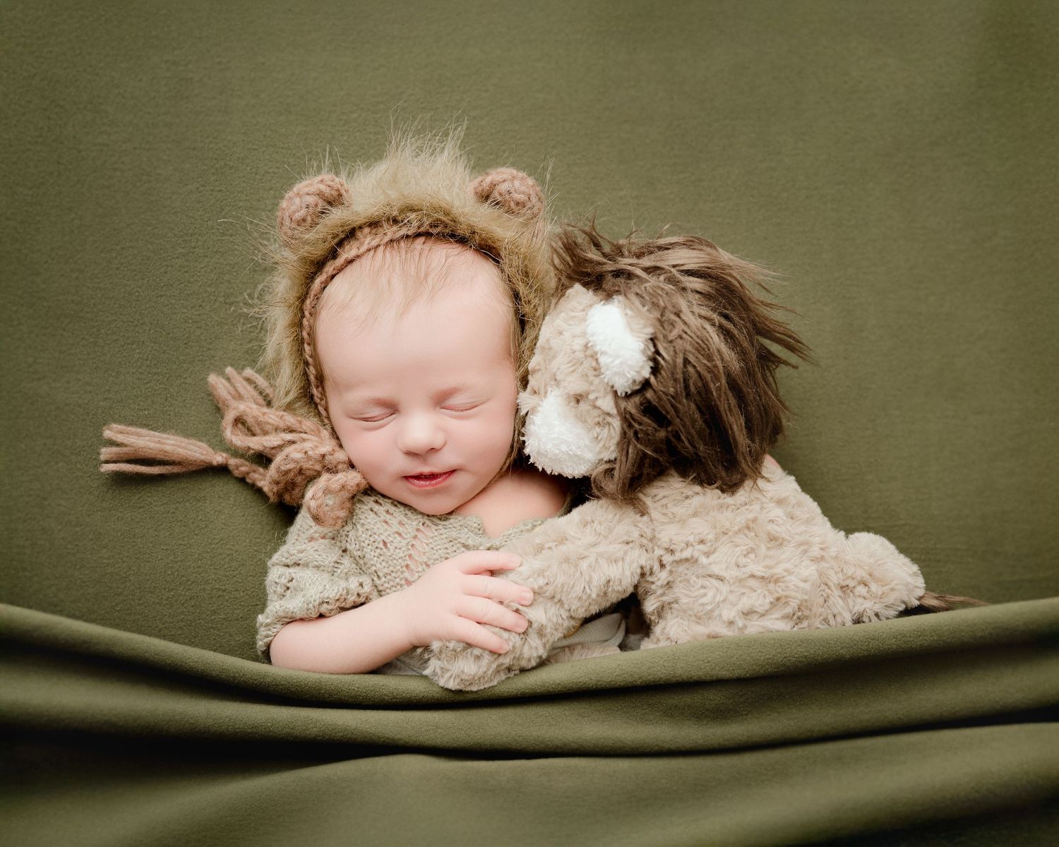 St Cloud newborn Photos of Sleeping newborn wearing a lion bonnet cuddles a lion stuffed animal on a green fabric backdrop.