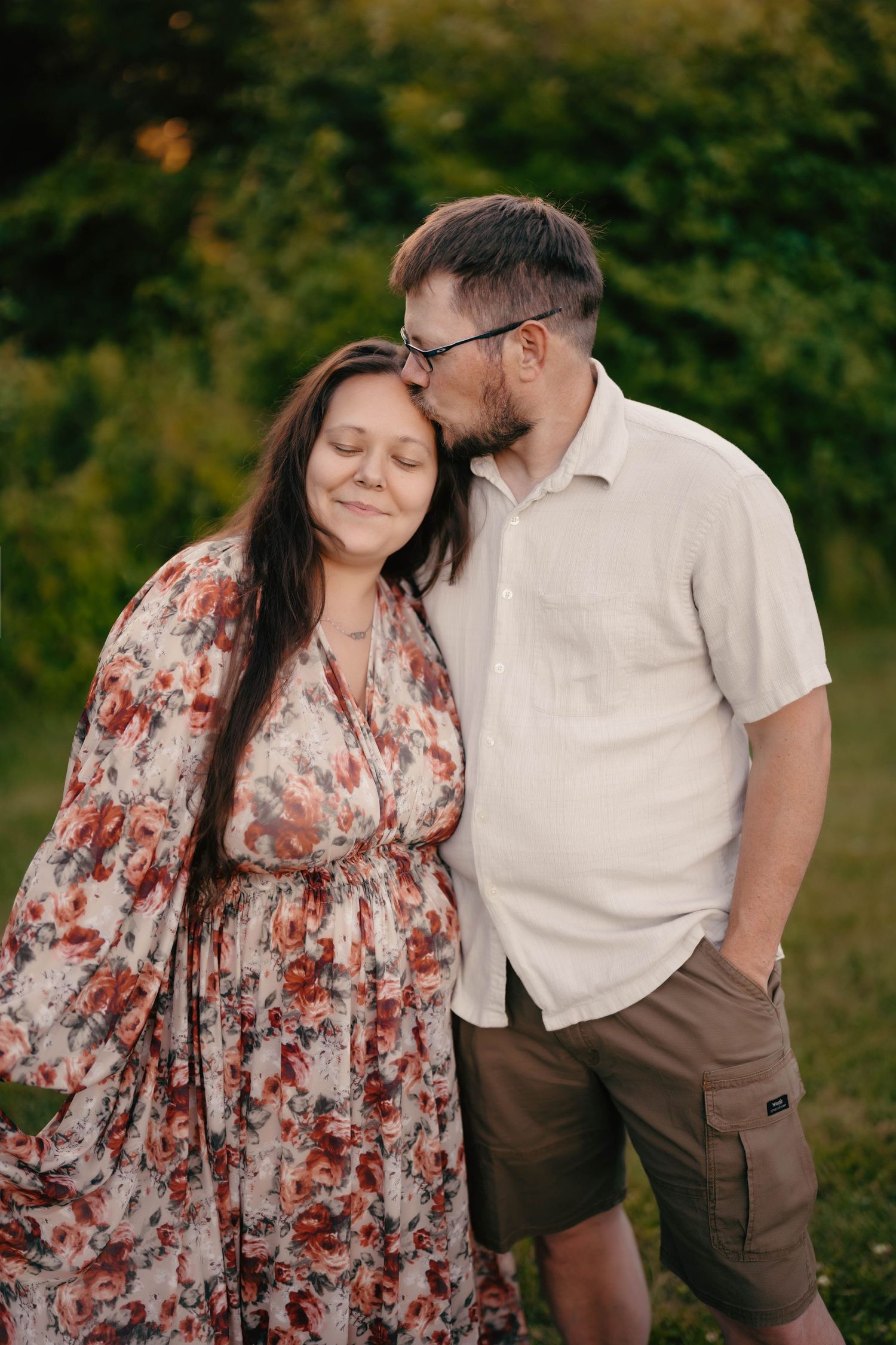 A couple shares a tender moment outdoors, man kissing woman's temple as she smiles in a floral dress.