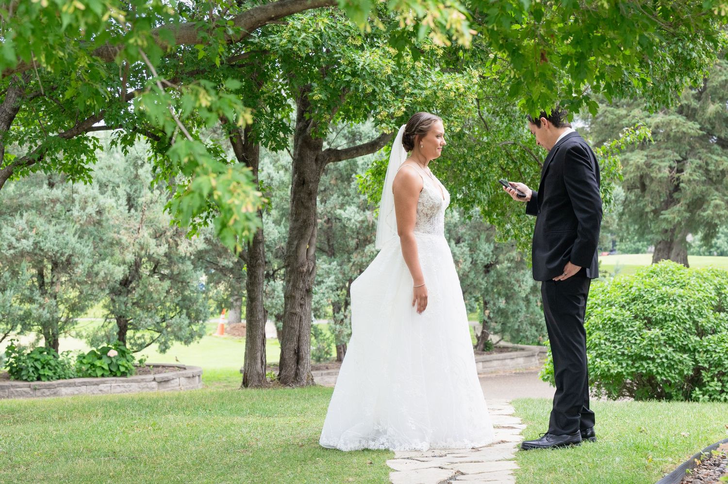 Bride in white gown and veil stands behind groom in black suit during outdoor first look ceremony.