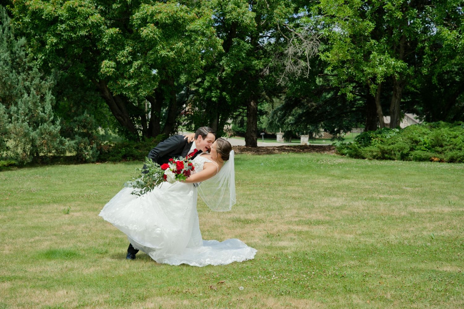 Groom dips and kisses bride in white wedding gown and veil on a green lawn surrounded by trees.