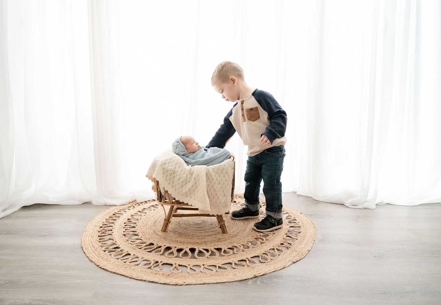 Minnesota Best newborn photographer -  a circular rattan rug is shown with a newborn and big brother on a grey floor against sheer curtains.