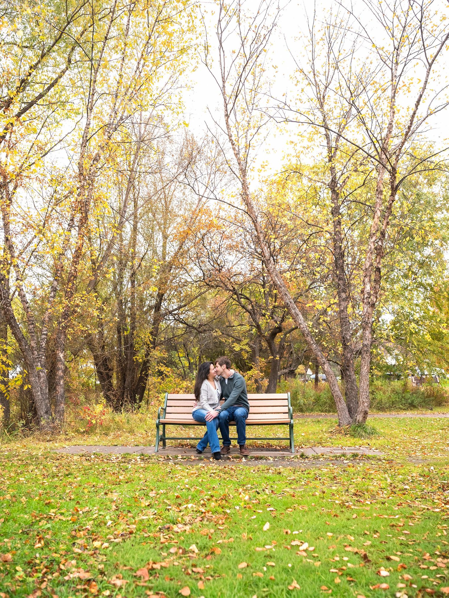 In central Minnesota a Couple sitting on wooden benches in a park surrounded by autumn trees with yellow leaves scattered on green grass.