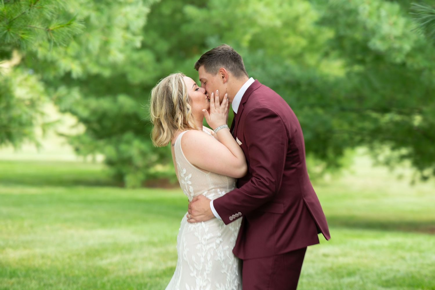 Romantic kiss between newlyweds in burgundy and white attire against lush green backdrop.