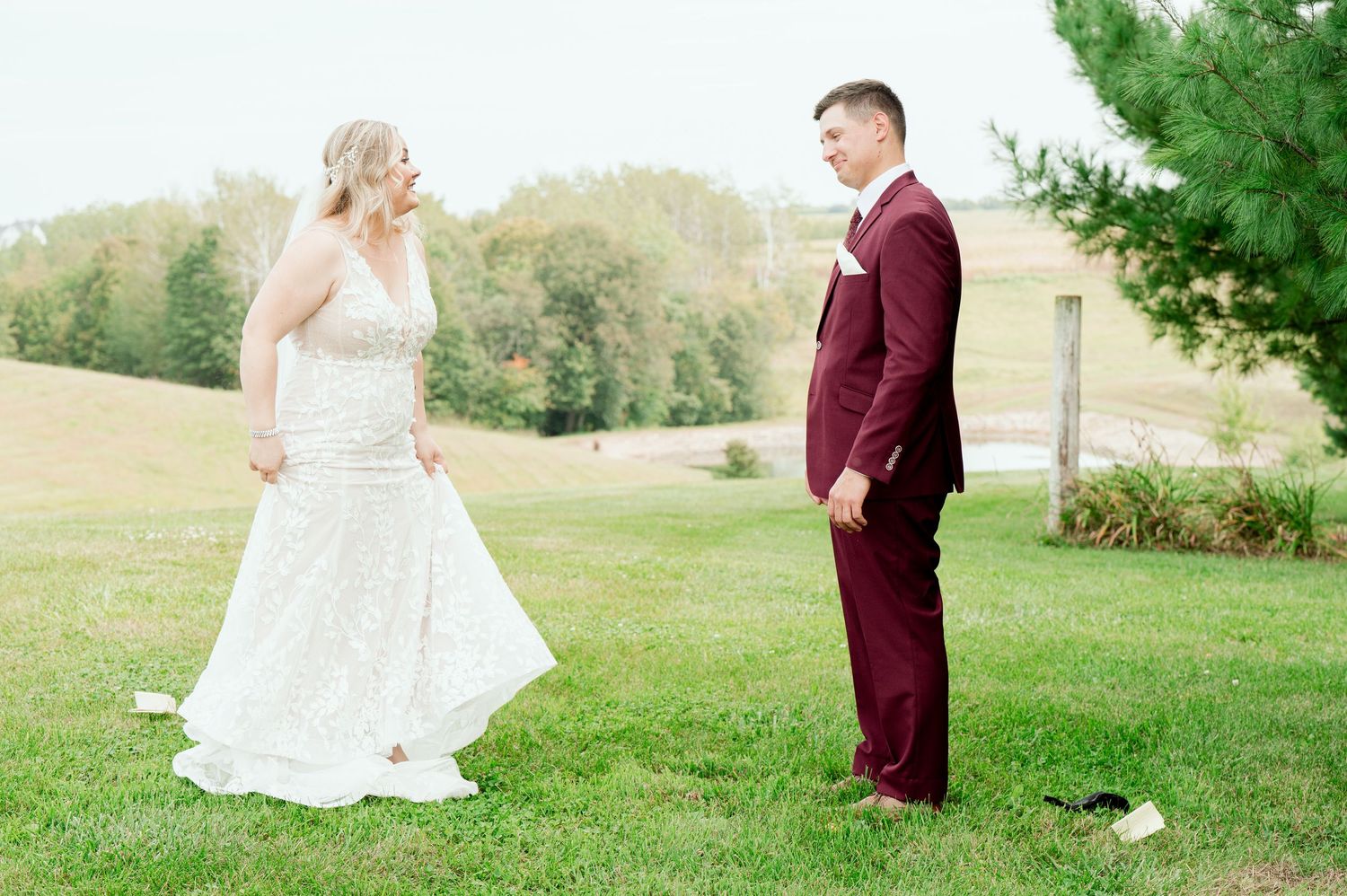 A bride in a flowing white gown faces her groom wearing a burgundy suit on a grassy hillside.