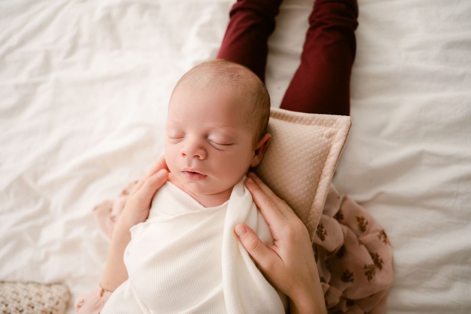 A peaceful sleeping newborn baby swaddled in white fabric lies on a cream-colored blanket.