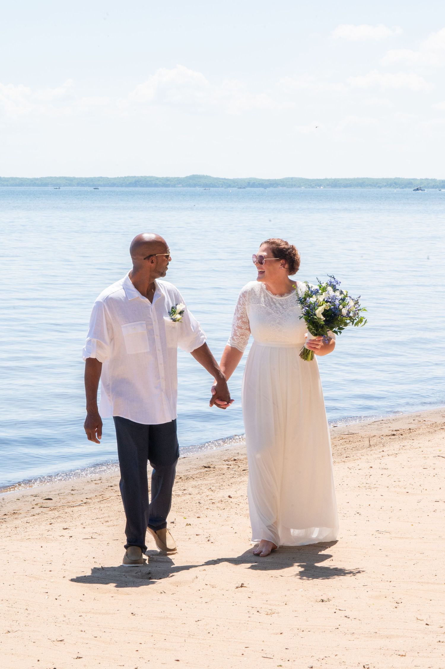 Newlywed couple holding hands walking on a sunny beach, bride in white gown with bouquet, groom in white shirt.