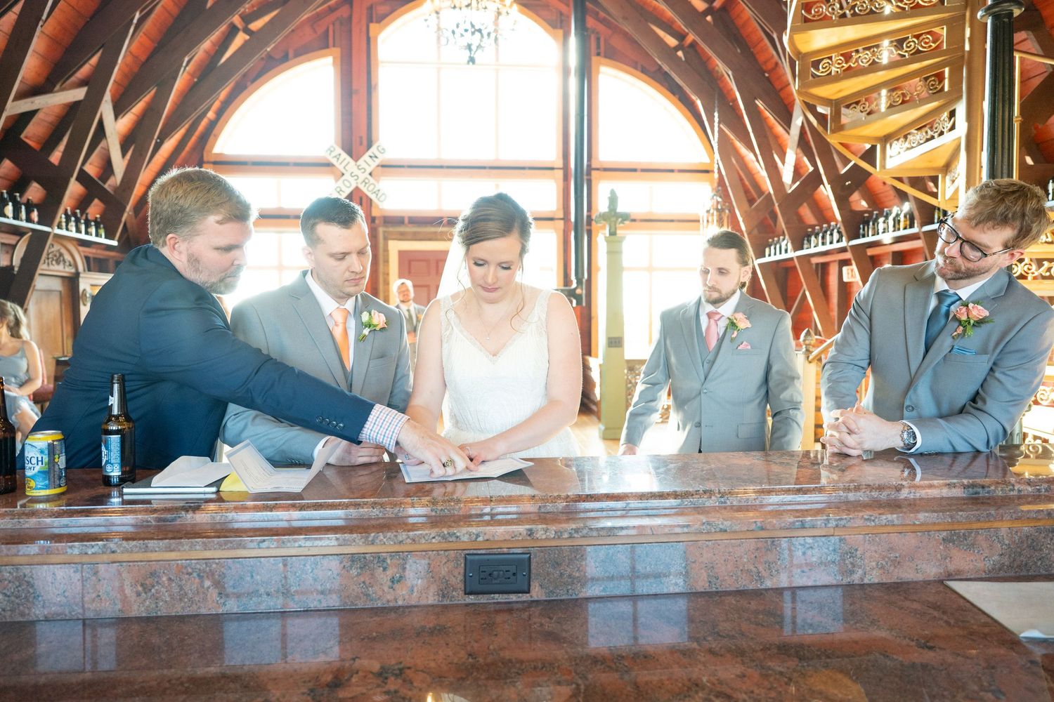 Wedding party gathers around a bar counter reviewing documents during a rustic venue reception ceremony.