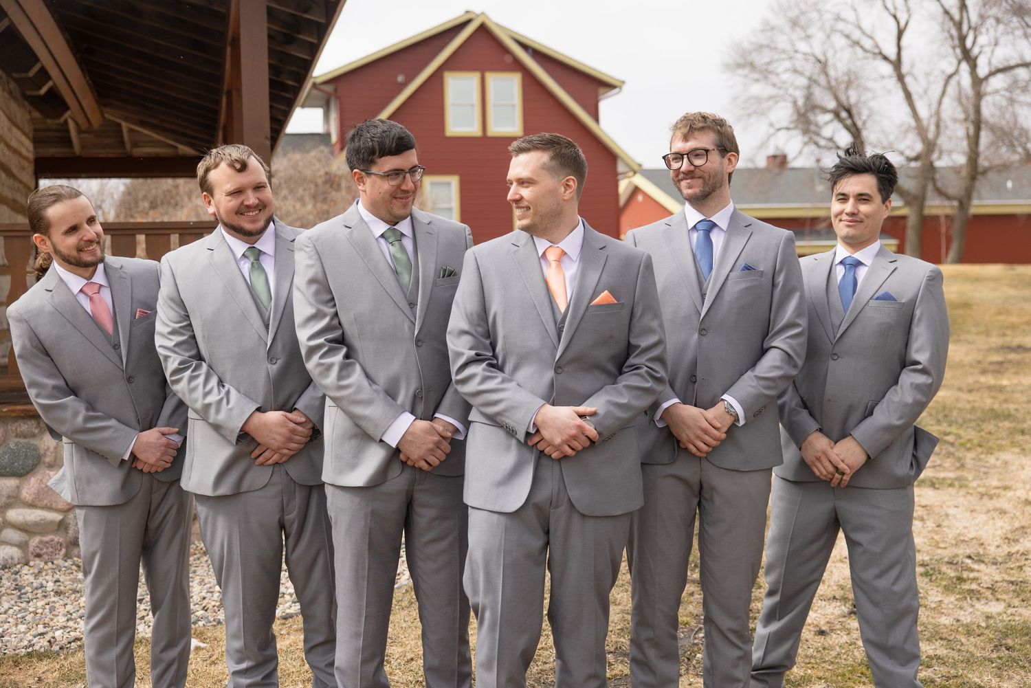 Groom and groomsmen posing in matching gray suits with peach and blue ties outside red barn venue.