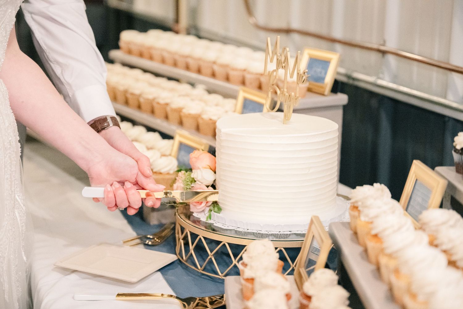 Bride and groom cut elegant white textured wedding cake surrounded by rows of decorated cupcakes.