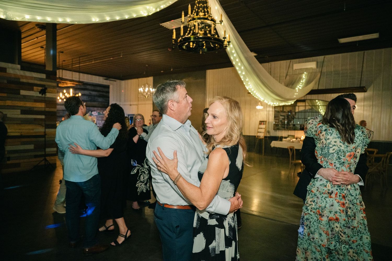 Guests dancing together at a wedding reception in a warmly lit venue decorated with draped fabric and chandeliers.