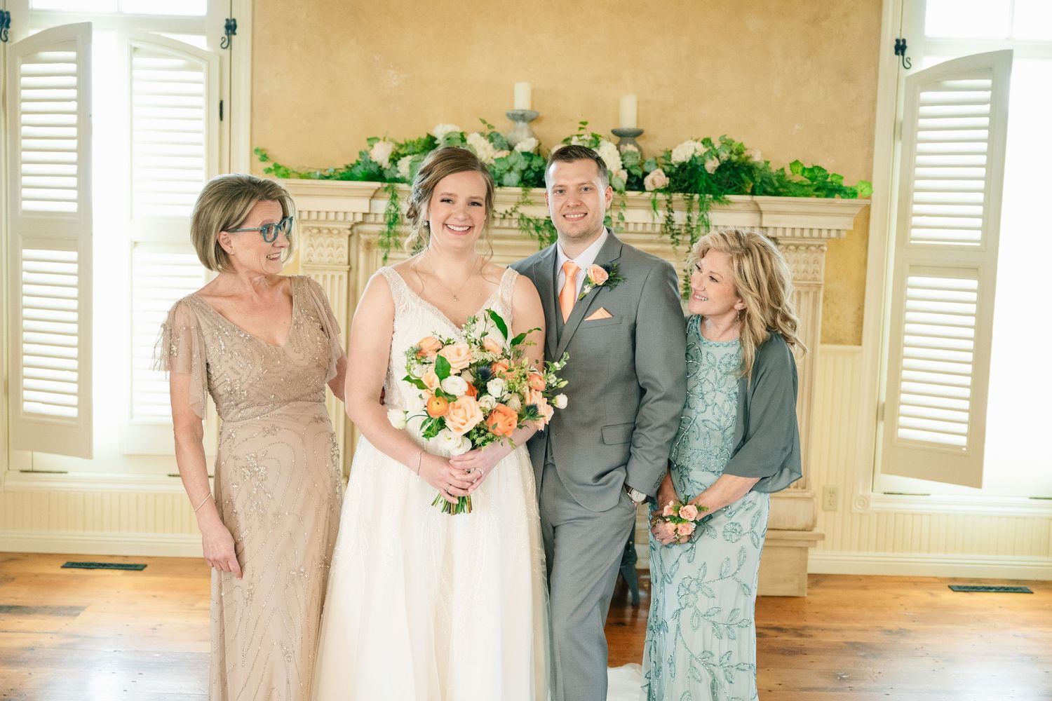 Bride and groom posing with two women near floral-decorated fireplace mantel in bright elegant room.