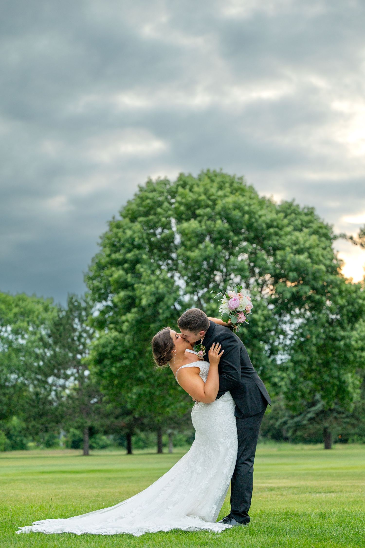 Bride and groom sharing romantic kiss on lush green lawn under dramatic cloudy sky.