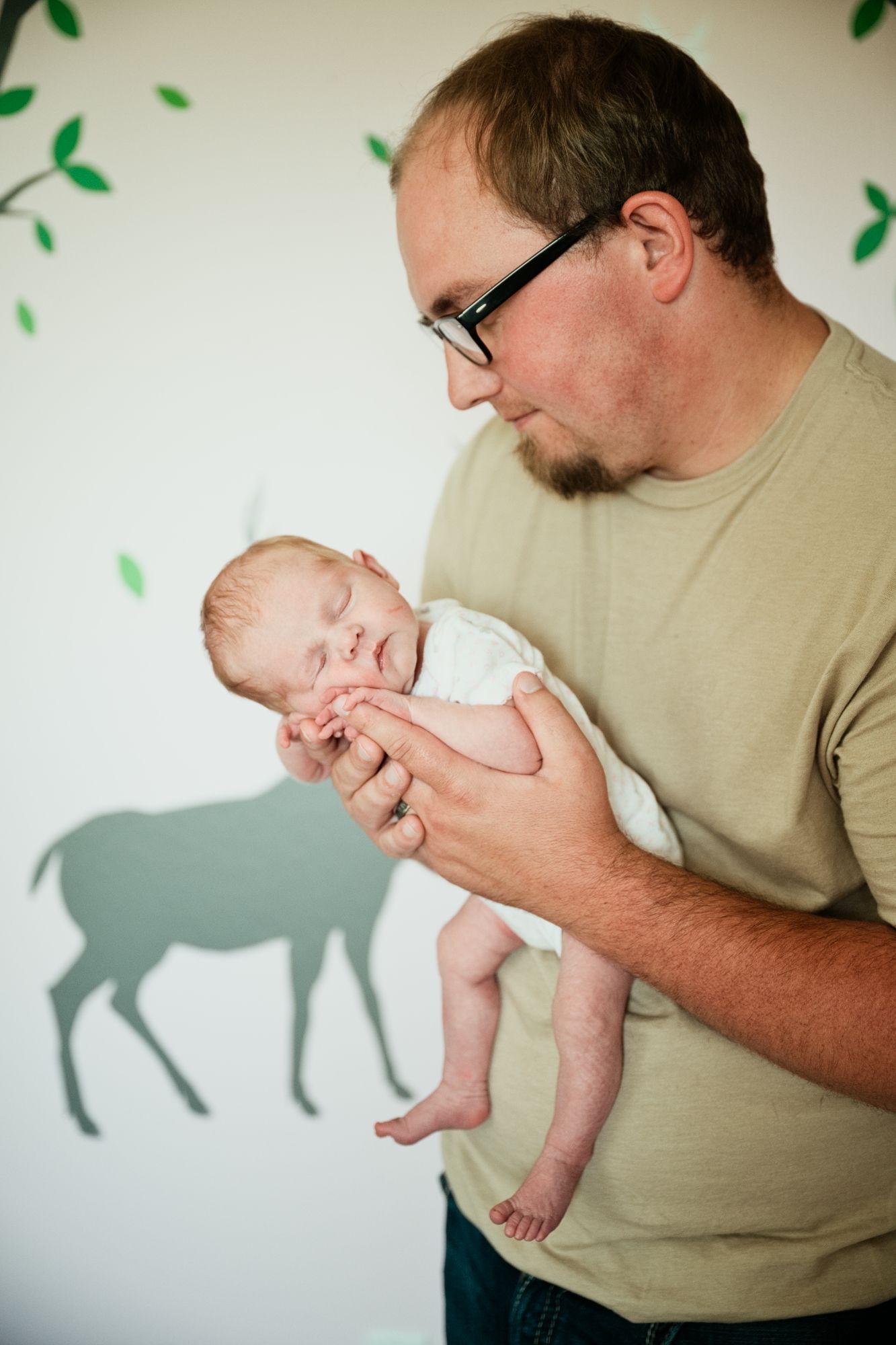 Father in glasses tenderly holds sleeping newborn baby against nursery wall with deer mural.