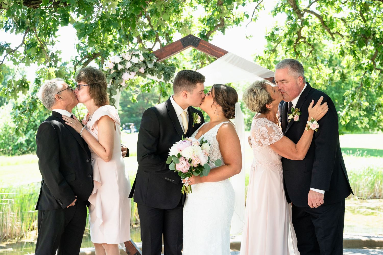 Wedding couple kisses at outdoor altar flanked by family members in formal attire amid lush green trees.